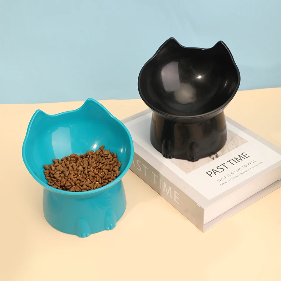 Deux gamelles surélevées en forme de tête de chat, une bleue avec croquettes et une noire, posées sur des livres