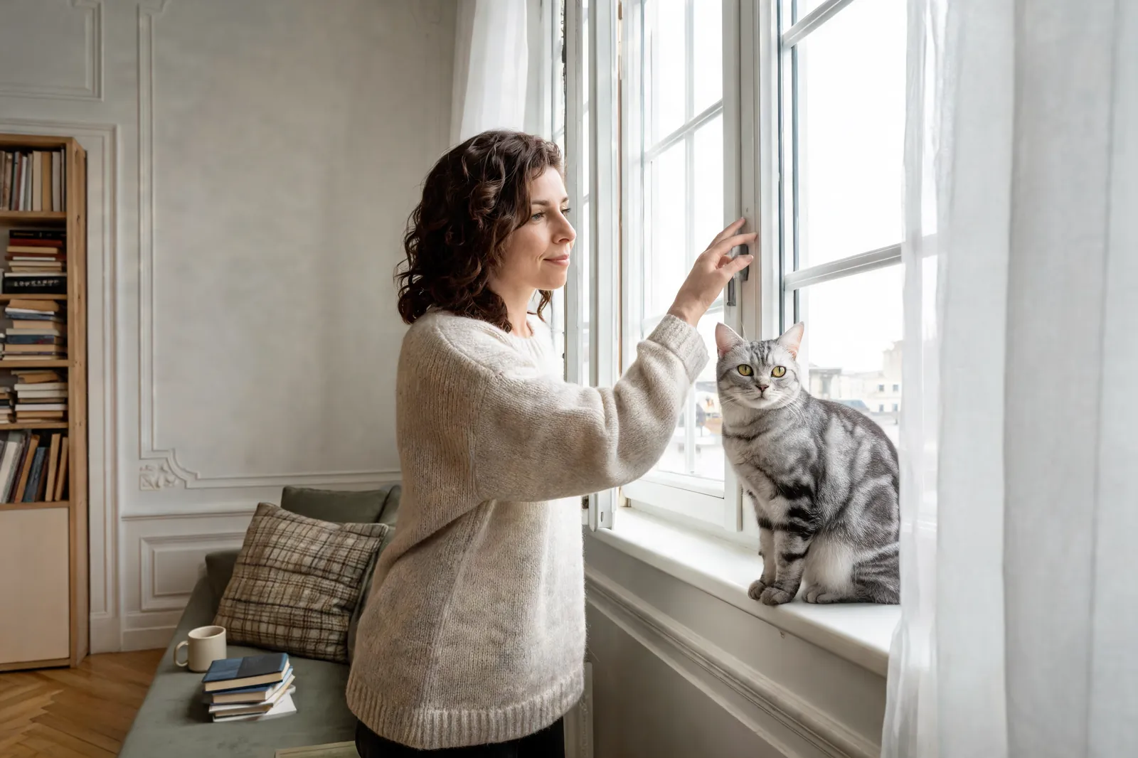 Une femme ajuste la poignée d’une fenêtre dans un salon lumineux, à côté d’un chat gris tigré assis sur le rebord